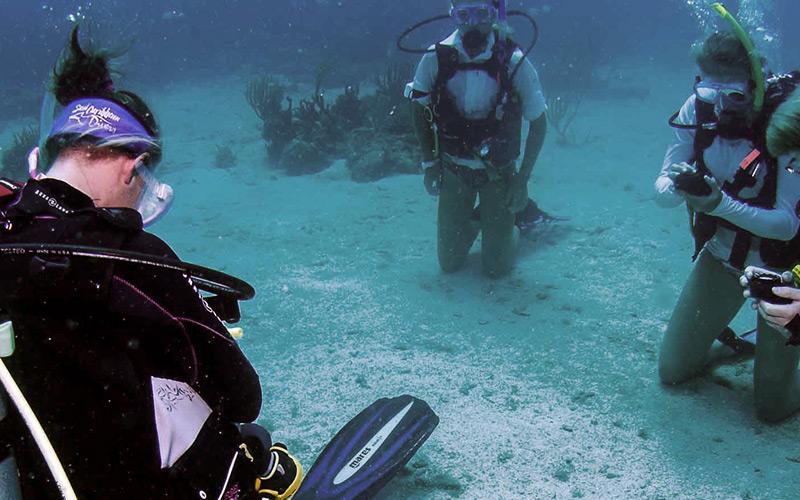 Students kneel on the ocean floor in scuba gear calibrating instruments while an instructor looks on.